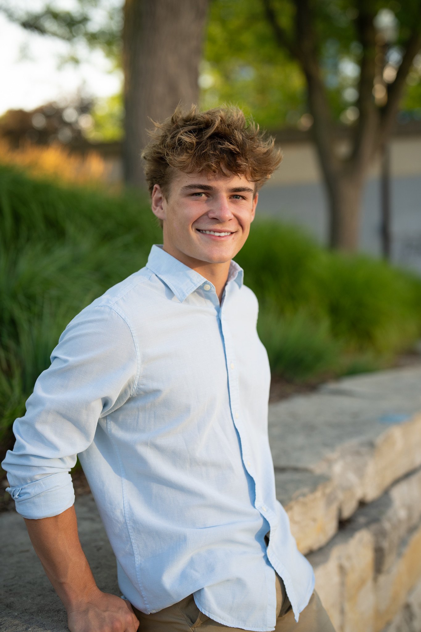 A young man with curly brown hair wearing a white shirt and tan pants, smiling and standing outdoors near trees and greenery.