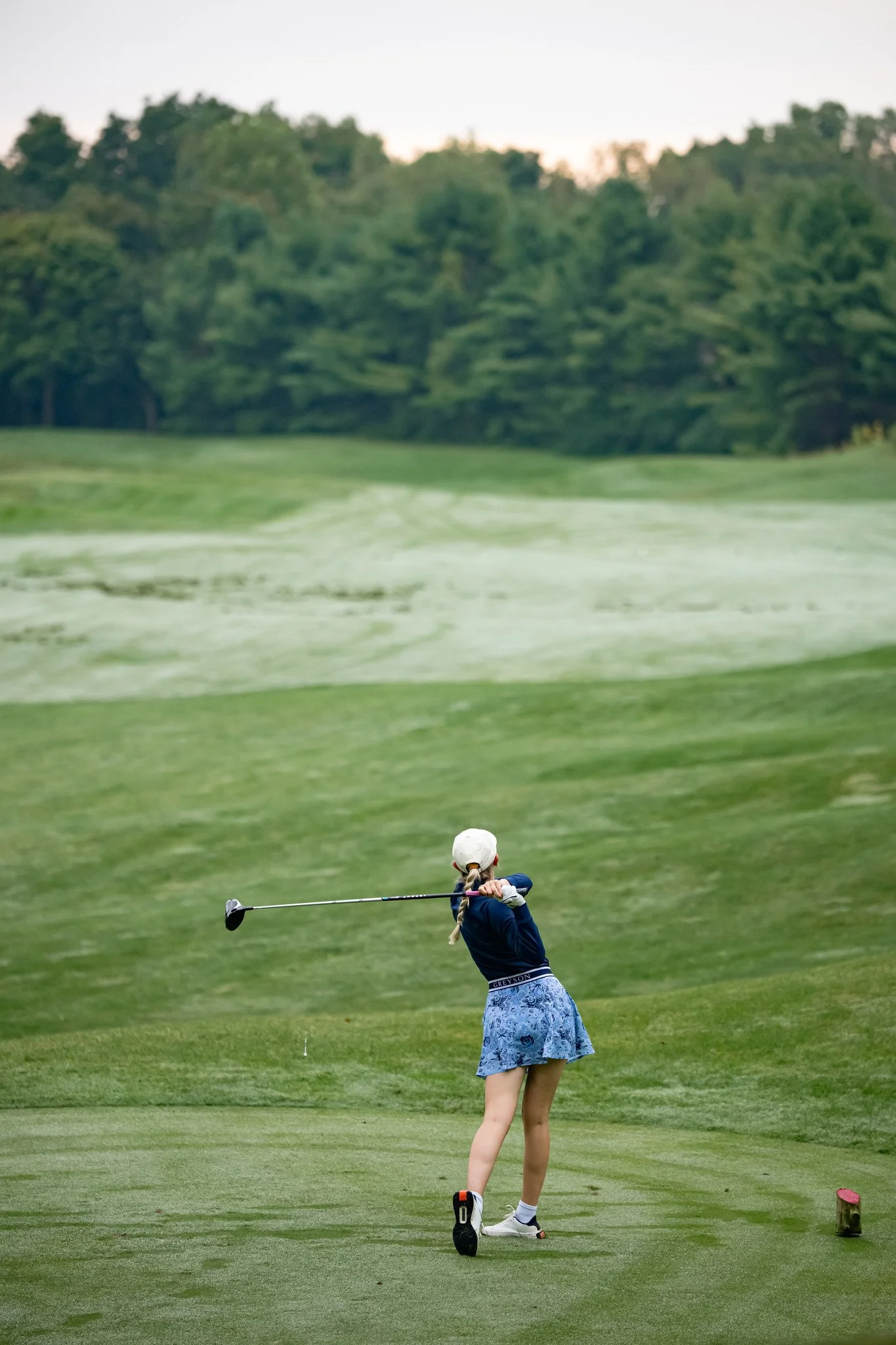 A young girl in a white cap and blue patterned skirt prepares to hit a golf ball on a golf course. She is standing on the green, with a pink tee marker nearby, and a wooded area in the background.