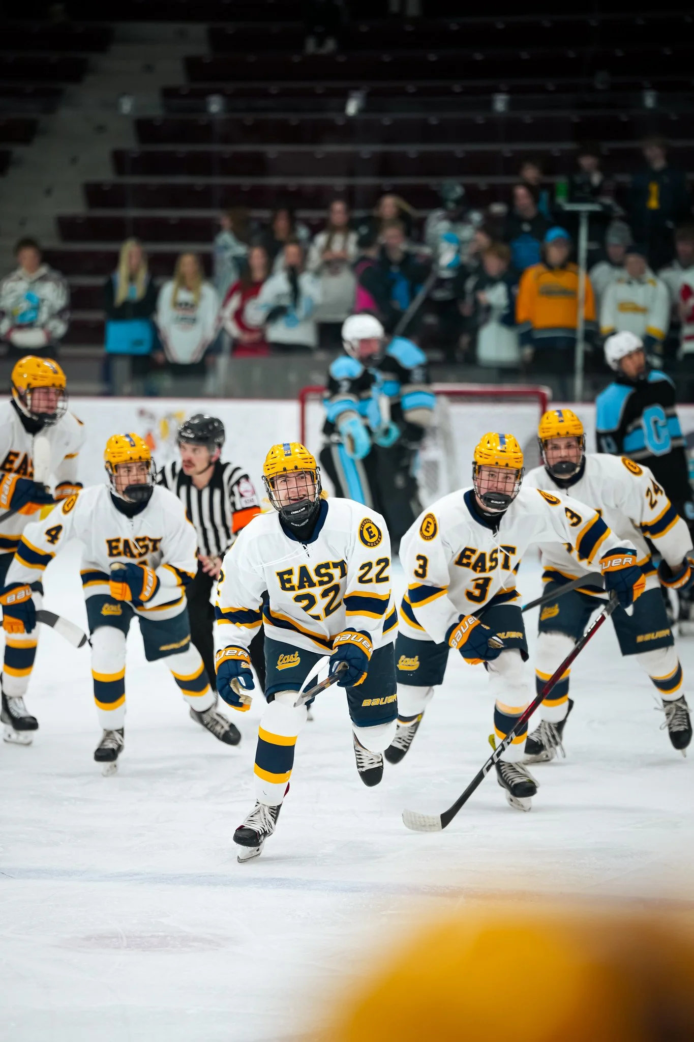 Ice hockey team in white and yellow jerseys skating on ice, with a referee nearby, and spectators in the background.