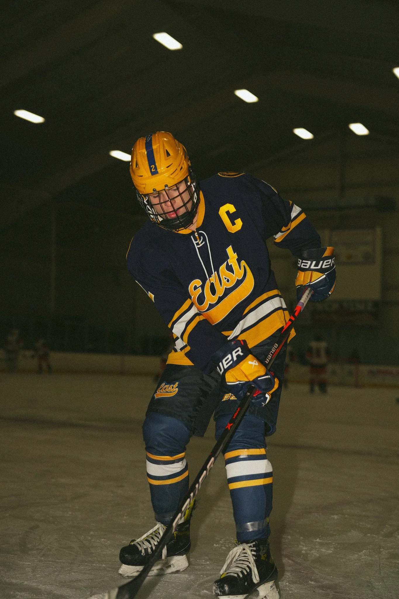 Ice hockey player in navy and yellow uniform with a captain's C, holding a hockey stick on ice rink.