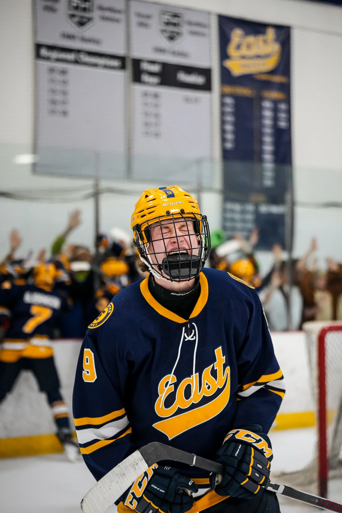 A young hockey player in a blue and yellow team jersey and yellow helmet is smiling on ice, with other players and scoreboard in the background.