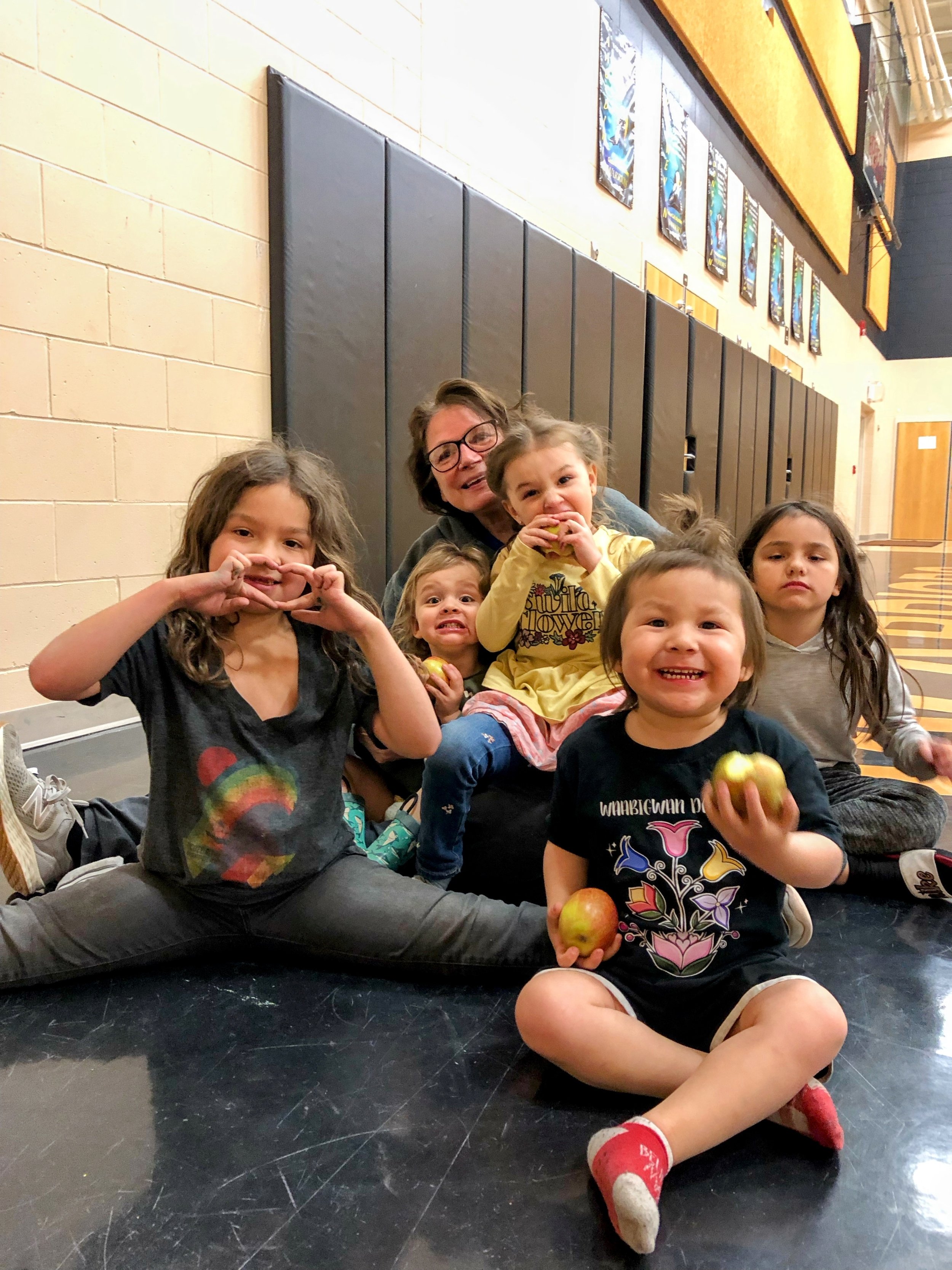 indigenous caregiver with glasses sits on the floor indoors surrounded by five young children making playful faces and posing for the camera, including a smiling toddler seated cross-legged in the foreground holding fresh apples.