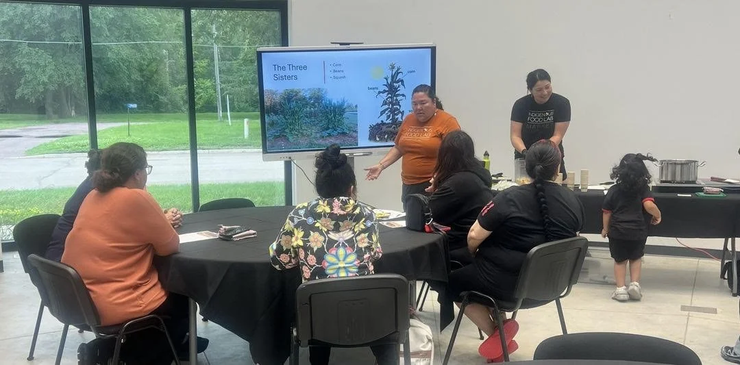 An Indigenous woman leads a community workshop about 'The Three Sisters' (corn, beans, and squash) using a digital display. Participants sit at a round table while another woman prepares food at a side station in a bright, modern room.