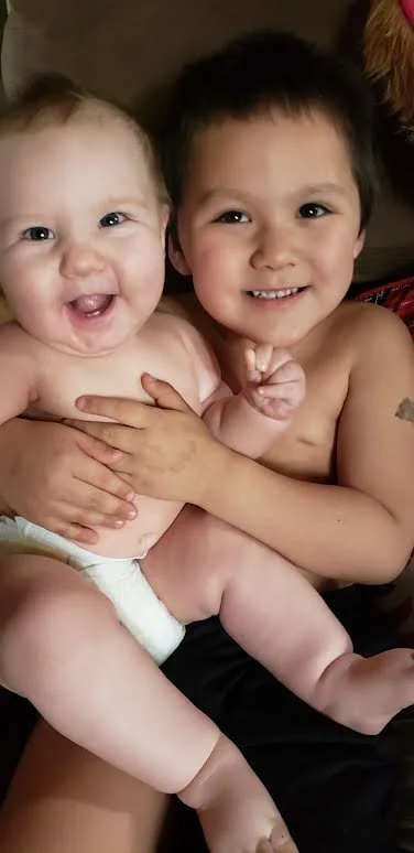 A close-up portrait of two young siblings or children sitting together; a smiling young boy has his arms wrapped gently around a laughing, blue-eyed baby wearing a white diaper.
