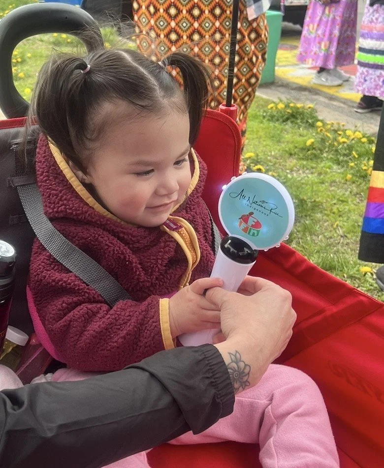 A young toddler with pigtails wearing a warm burgundy fleece and pink pants sits inside a red wagon outdoors, looking down and smiling as an adult holds out a small light-up wand for her