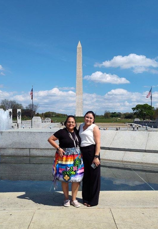 Two women smiling and posing at the World War II Memorial, with the Washington Monument in the background. One woman wears a vibrant, multi-colored Indigenous ribbon skirt with floral embroidery.