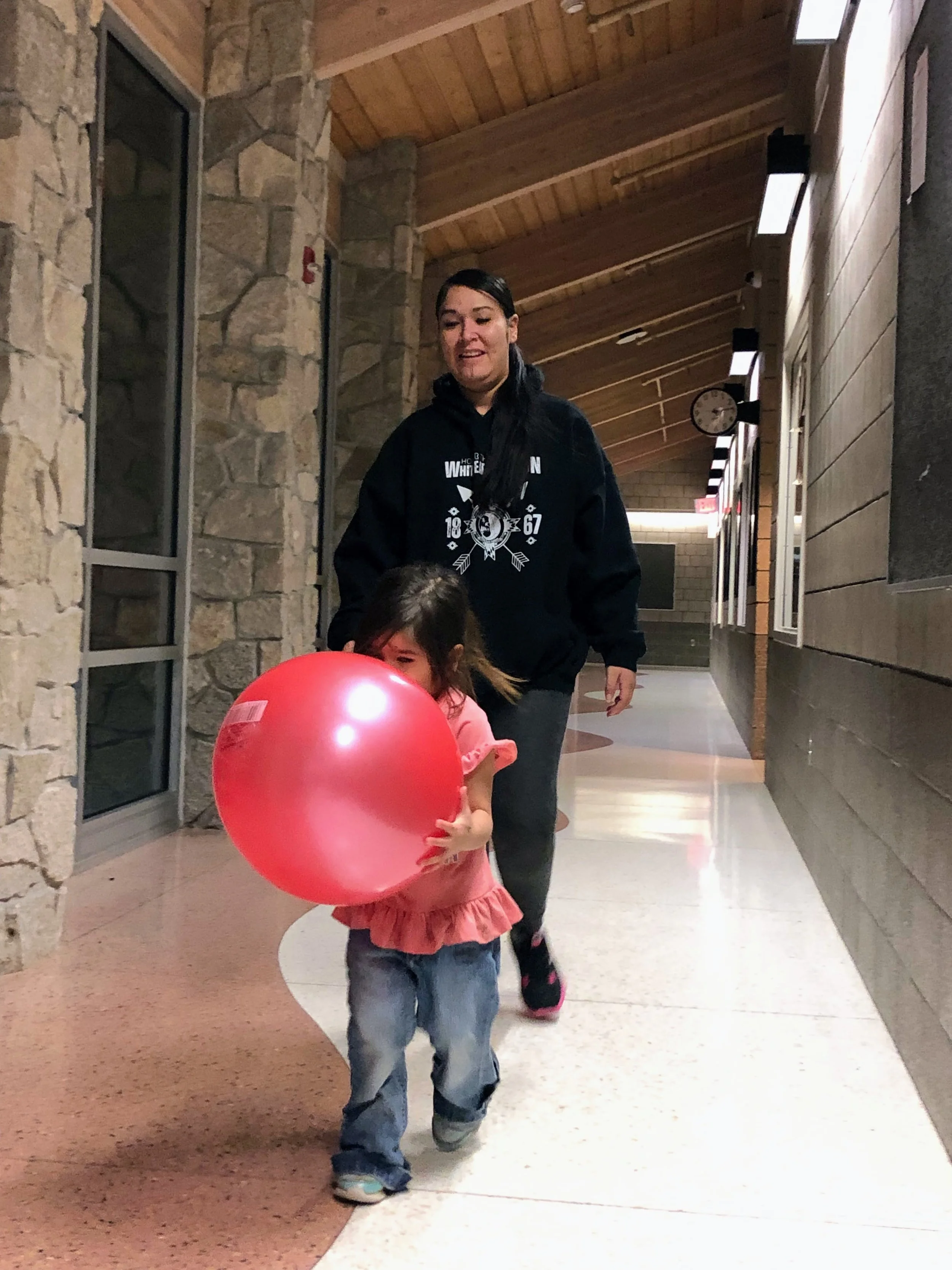 A young child in a pink shirt and jeans carries a large red ball down a bright indoor hallway, followed by a woman in a black hoodie