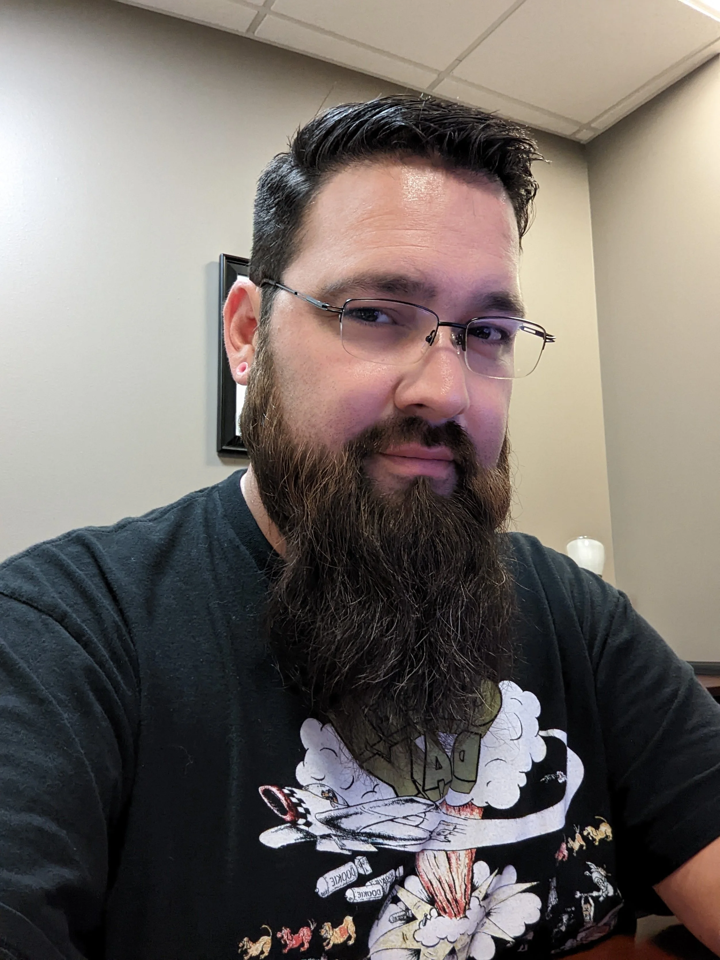 A close-up headshot portrait of the IT Specialist, a man with dark hair, glasses, and a long dark beard, looking directly at the camera in a modern office environment.