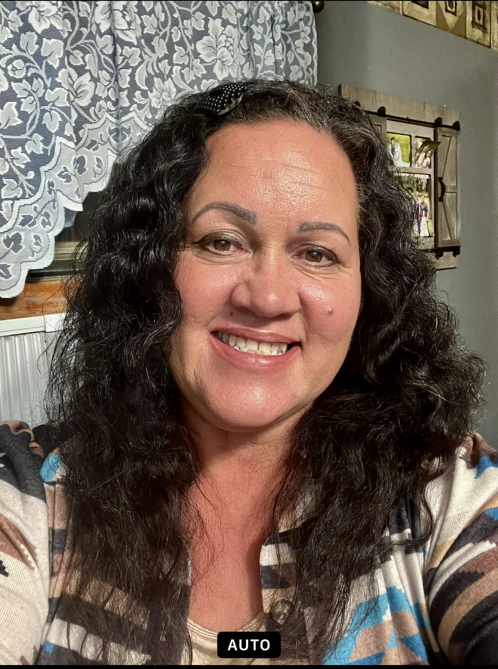 A close-up headshot portrait of the Finance Officer, a smiling woman with long dark wavy hair wearing a patterned top, in a home setting with decorative wall accents in the background.
