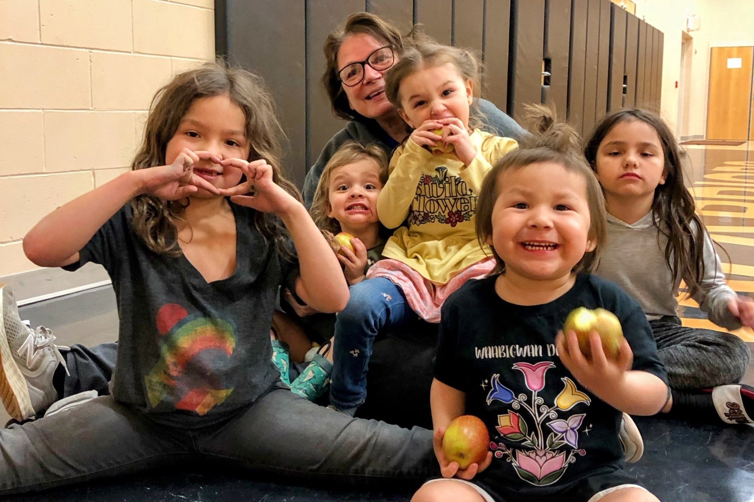 A heartwarming group photo of six Indigenous children and one adult Indigenous woman smiling and laughing together on an indoor basketball court. Some of the children are holding and eating apples, and one girl is making a heart shape with her hands
