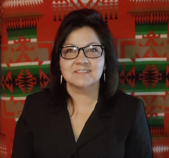 A headshot portrait of the CEO, an Indigenous woman wearing rectangular glasses and a black blazer, posed in front of a vibrant red, green, and earth-toned geometric woven textile backdrop.