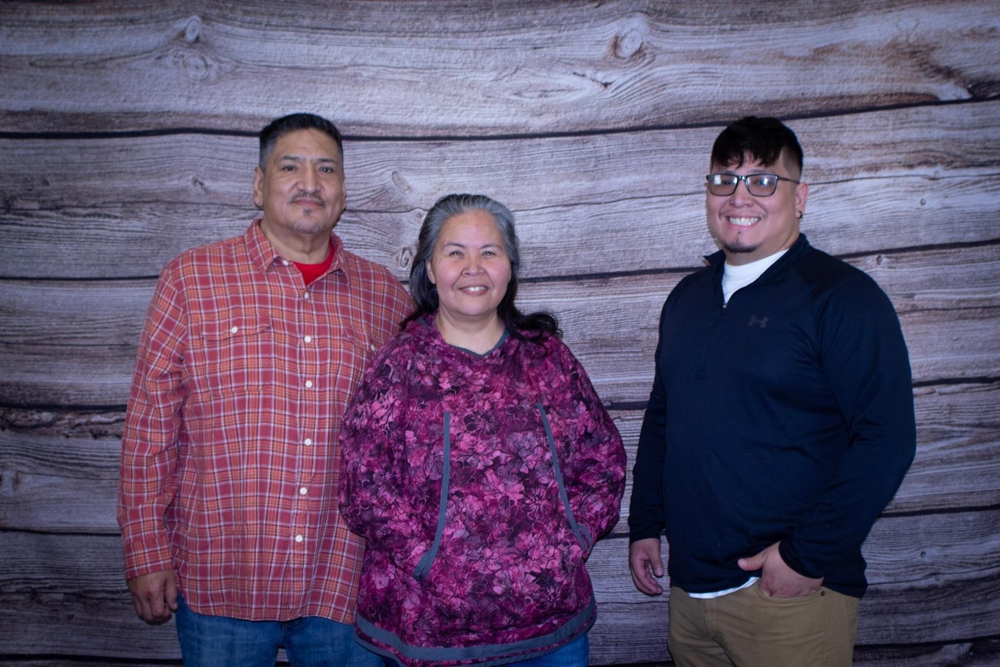 IPLI participants standing side-by-side in front of a faux wood wall; a man in an orange plaid button-down on the left, a woman in a purple patterned hoodie in the middle, and a smiling man wearing glasses and a dark long-sleeve shirt on the right