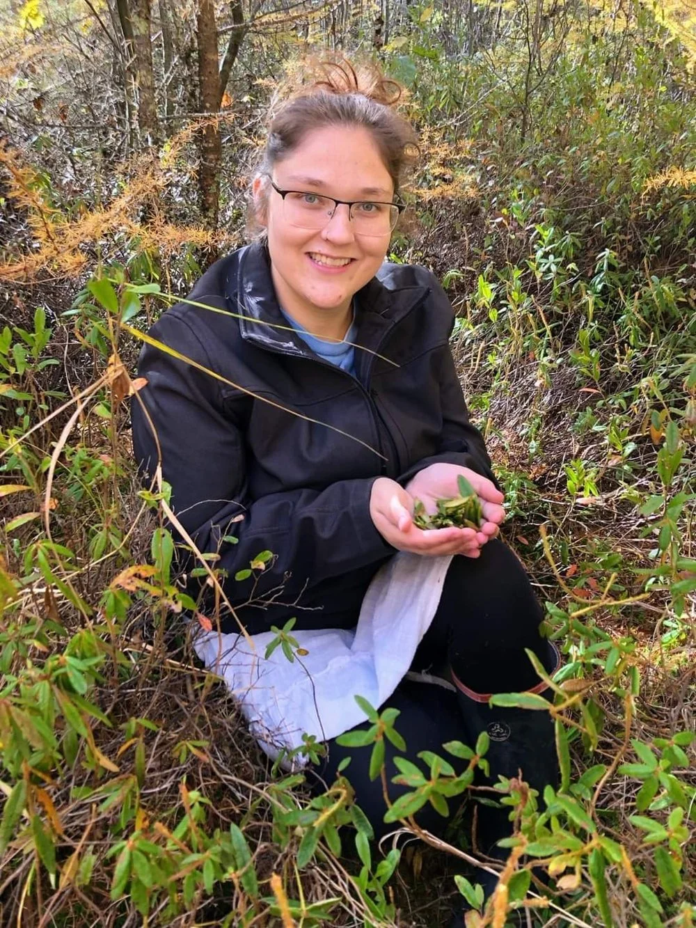 Outdoor portrait of Marlena wearing glasses and a black jacket, smiling as she kneels outdoors in a lush wooded area and cradles freshly gathered green plants in both hands.