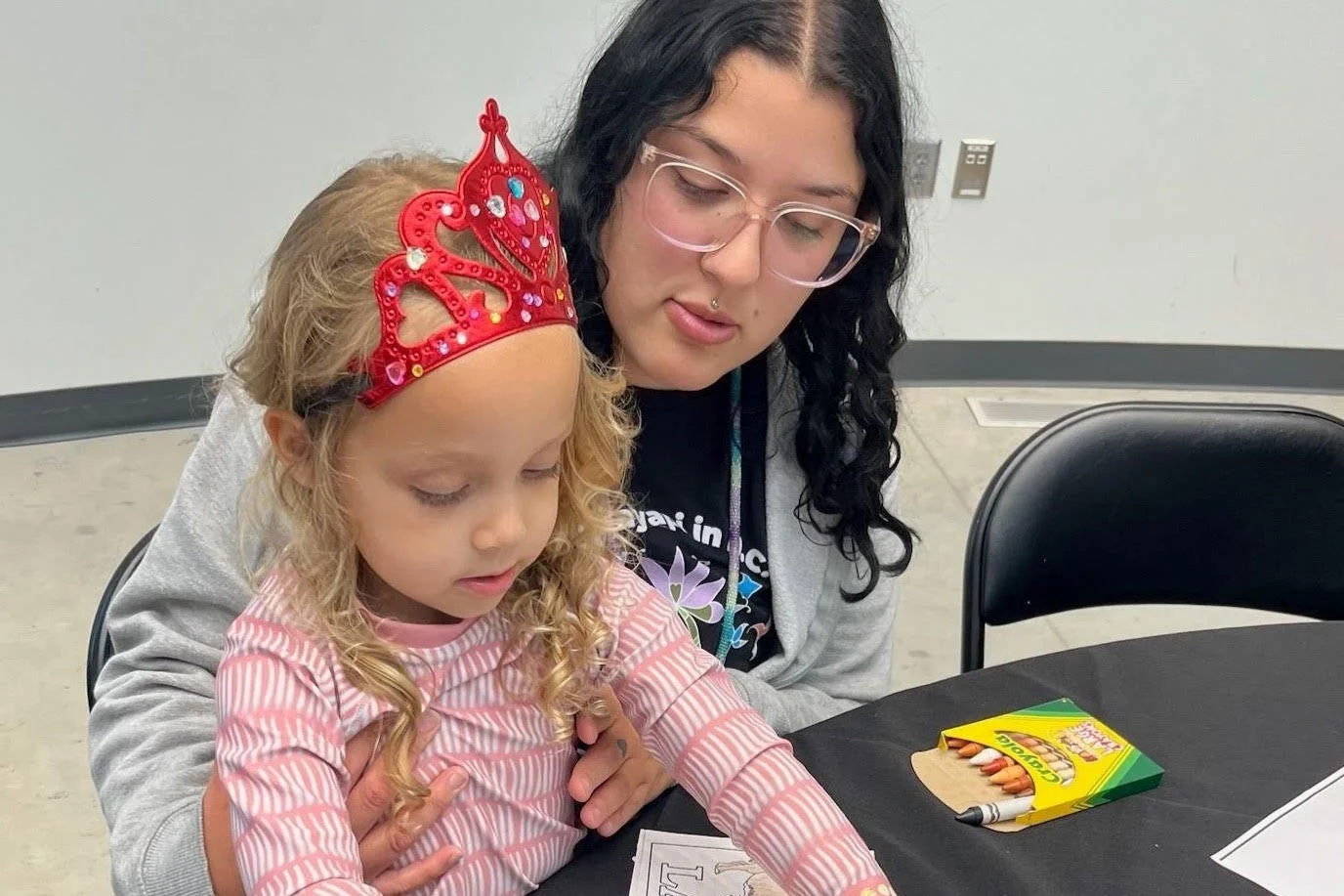 An Indigenous woman with long dark hair and glasses sits closely with a young girl wearing a bright red, jeweled princess tiara. They are both focused on a coloring page on a black table, with a box of crayons nearby.