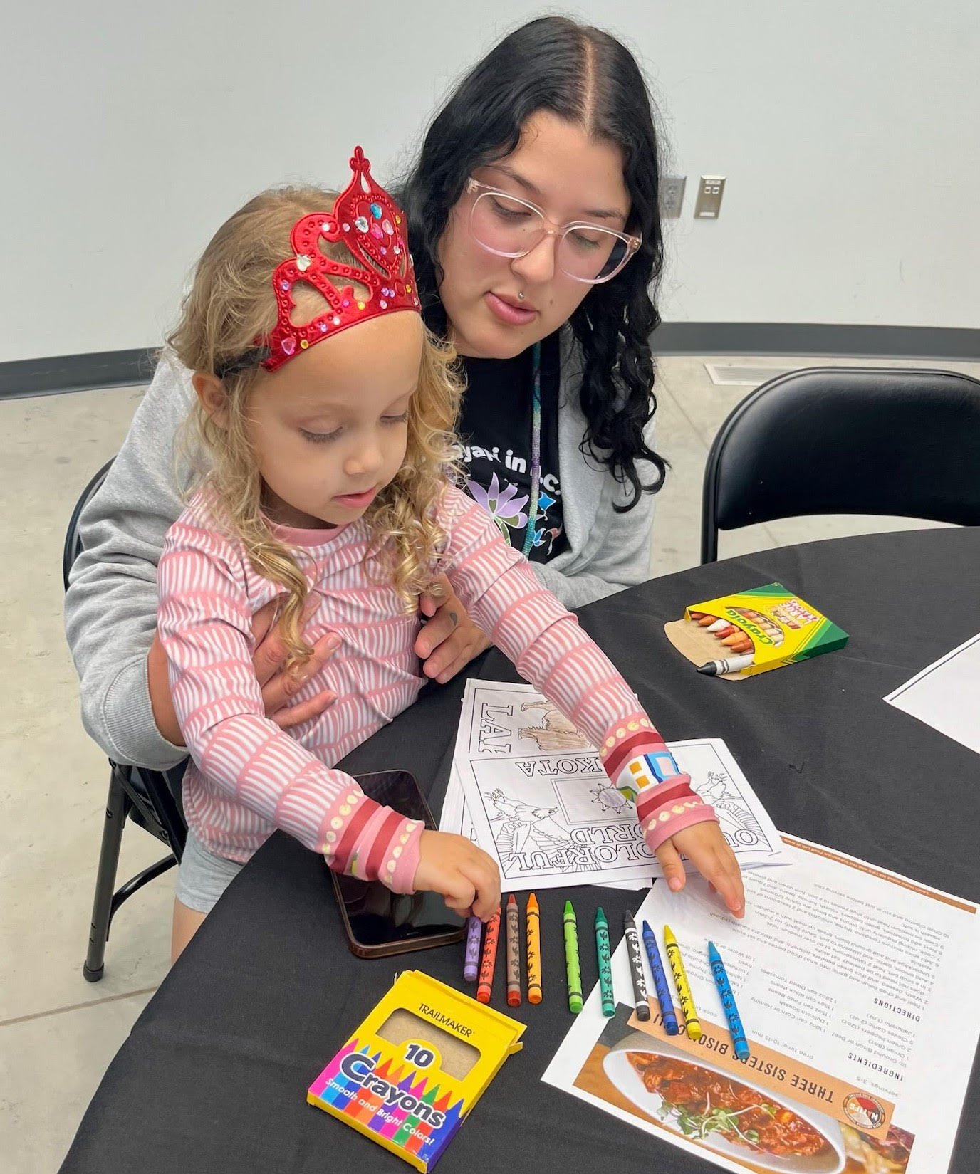 A young girl with blonde curls wearing a sparkly red princess tiara and a pink patterned long-sleeve shirt reaches for colorful crayons lined up on a black table beside coloring pages, with an adult woman wearing clear glasses leaning in beside her.