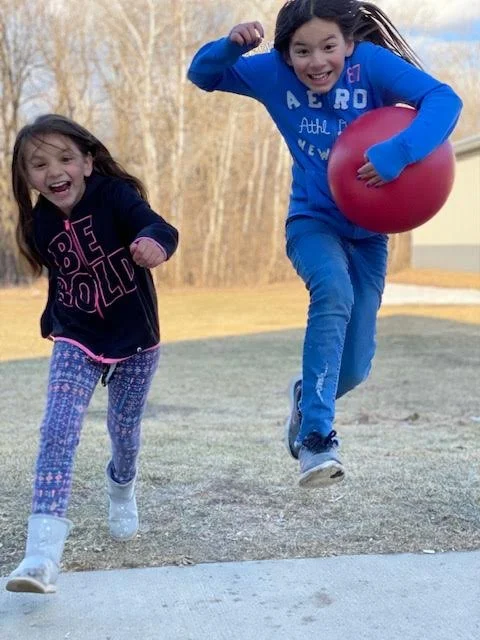 Two excited young girls running together across a grassy yard; a girl on the right wearing a bright blue outfit runs toward the camera carrying a large red ball, while a younger girl on the left in a black hoodie and purple leggings