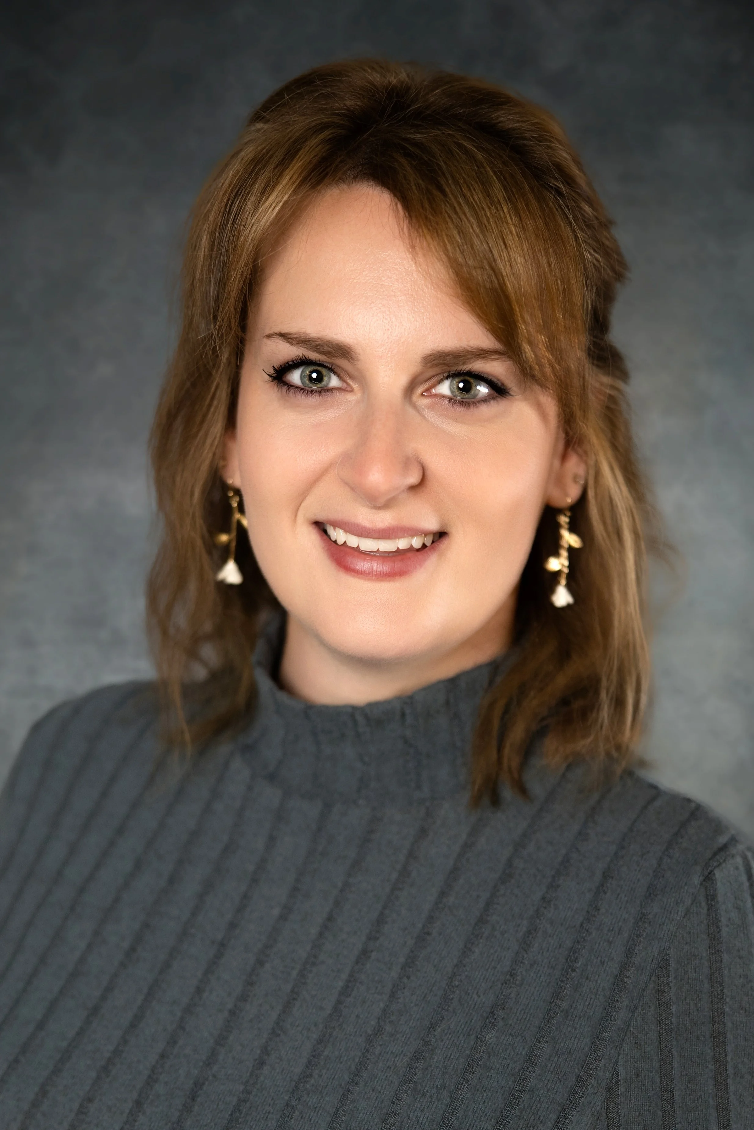 A professional headshot portrait of Candace Rastedt featuring shoulder-length brownish-blonde hair and dangling earrings, smiling warmly while wearing a charcoal gray ribbed turtleneck sweater in front of a textured gray studio background.