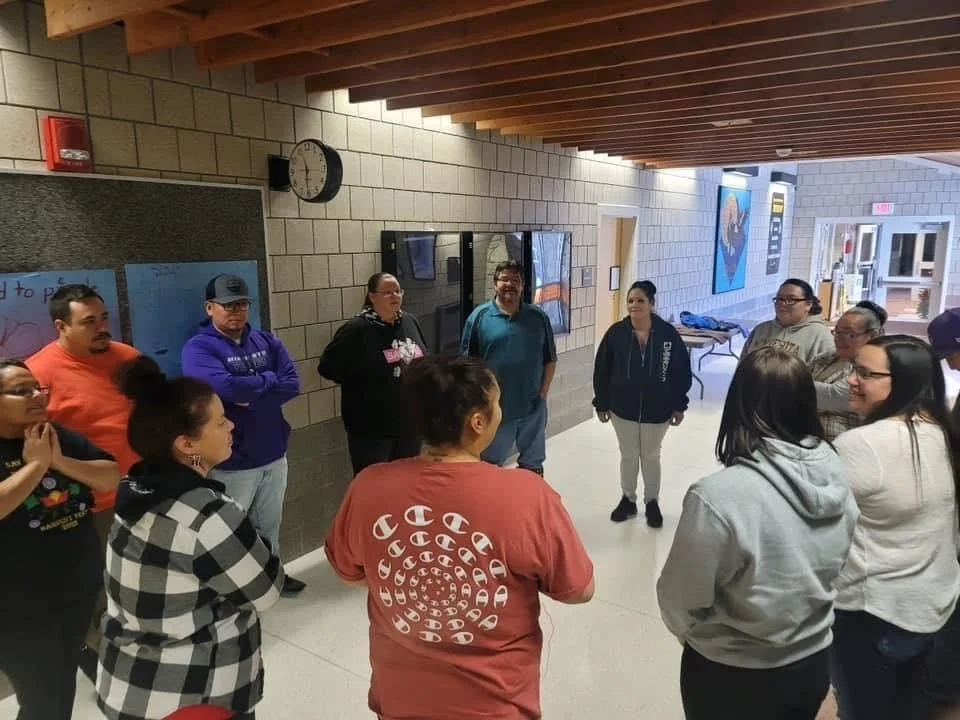 A wide indoor shot of an Indigenous Parent Leadership Initiative (IPLI) cohort standing in a circle in a modern hallway with exposed wood-beam ceilings and brick walls, engaged in a group conversation or debrief