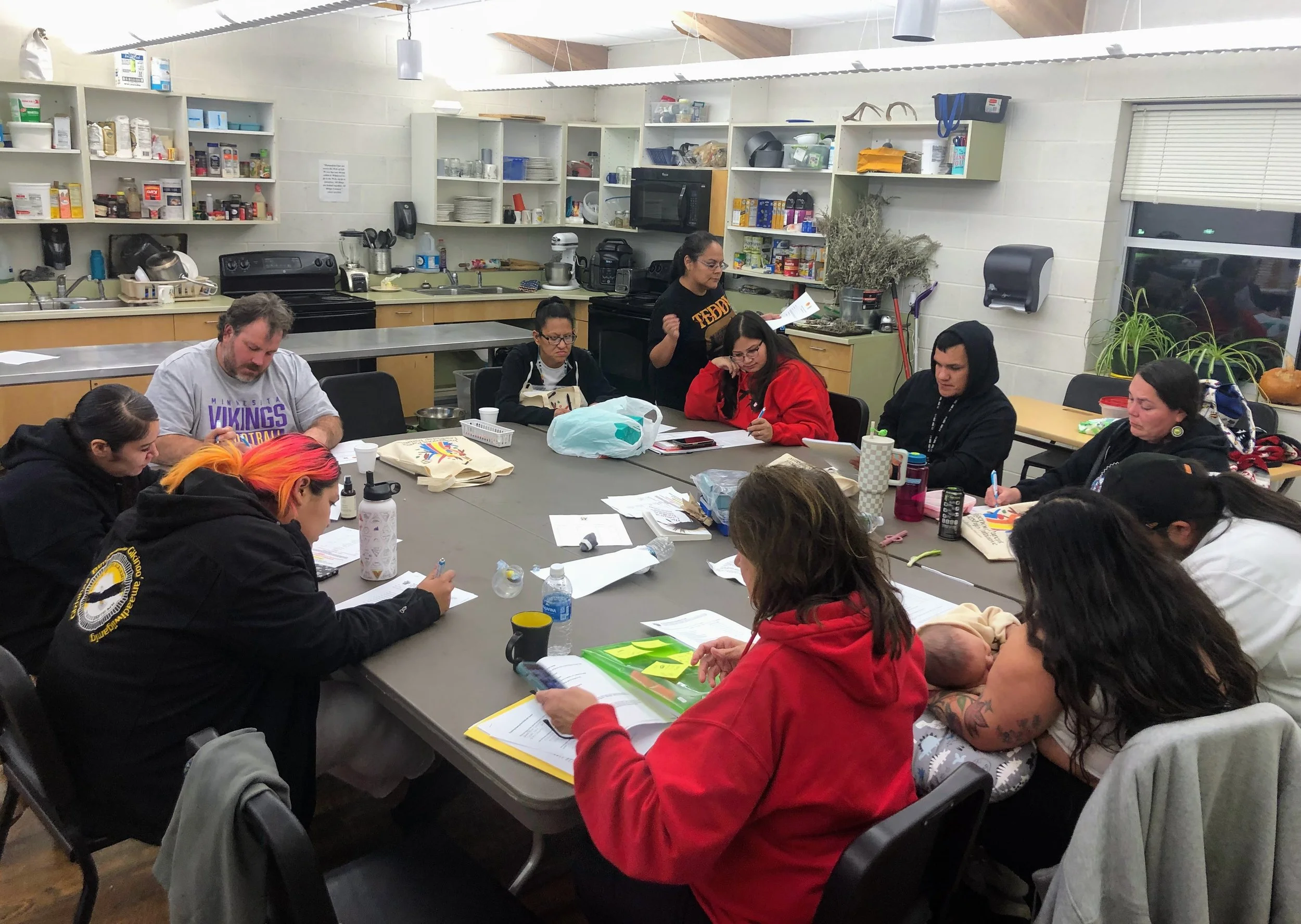 a group of Indigenous parents and community members seated around a large table working on papers and activities in a kitchen-style meeting room, beneath the title "Indigenous Parent Leadership Initiative."