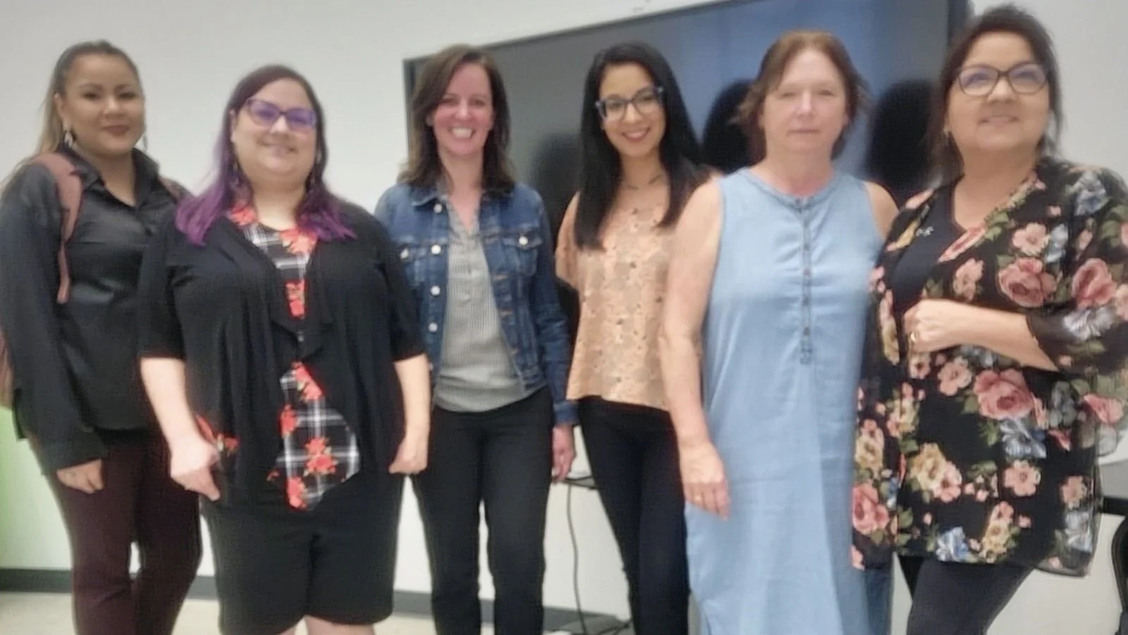 "A group portrait of six women from the First Children First Nation (FCFN) team standing together in an office setting. They are smiling and dressed in business-casual attire, including floral patterns, denim, and professional tops