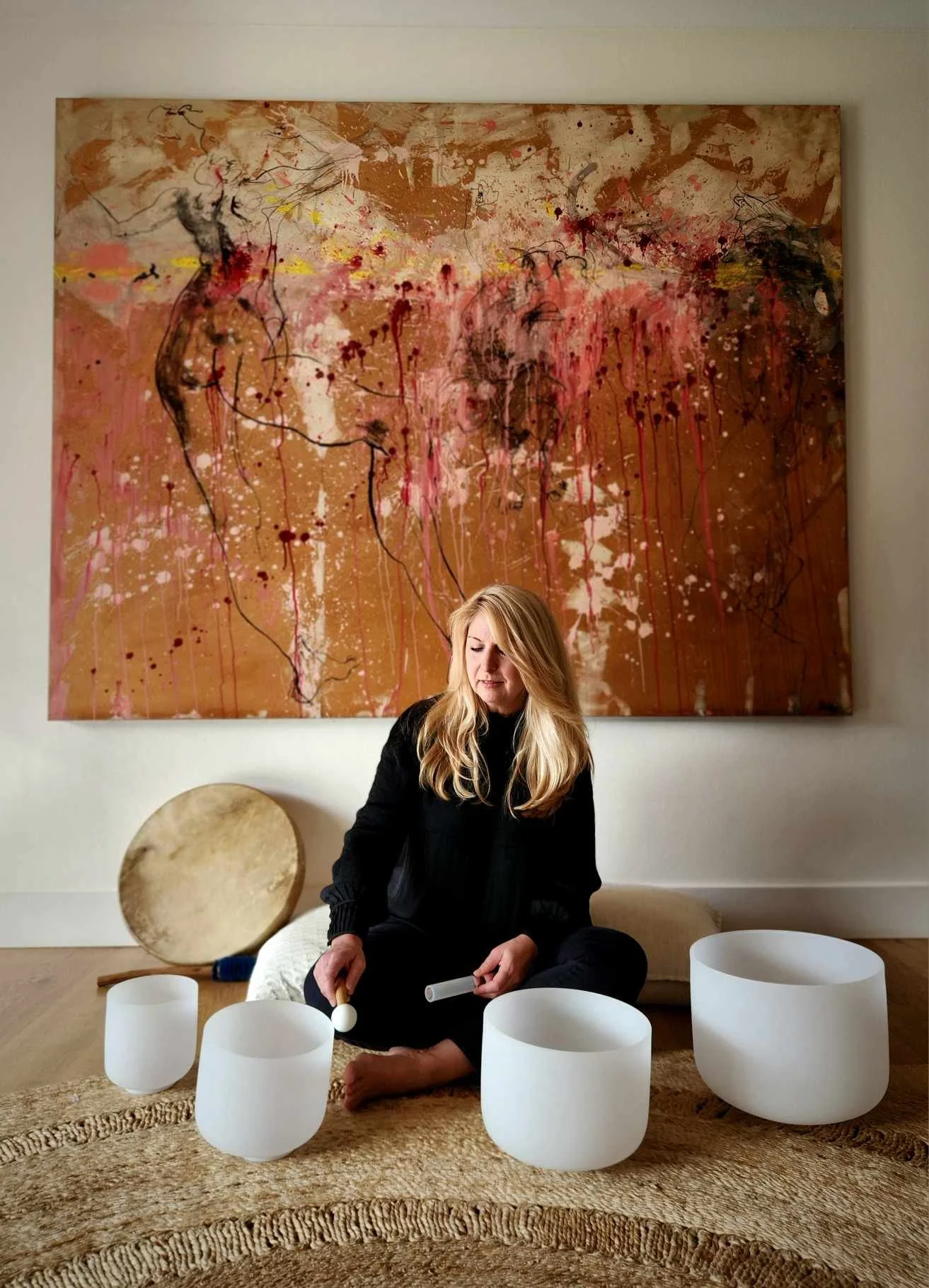 Amber Clements sitting cross-legged on a rug, holding a singing bowl and a mallet, surrounded by three more singing bowls, with a large abstract painting hanging on the wall behind her.