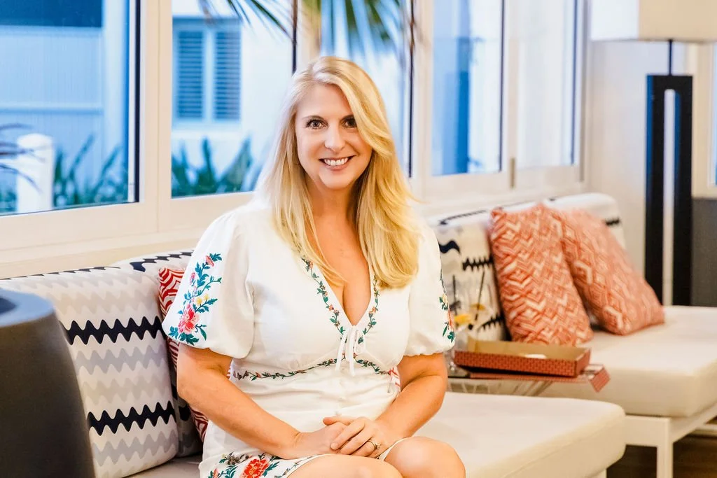 Amber Clements in a white embroidered dress sitting on a patterned sofa in a brightly lit room with large windows and red pillows.
