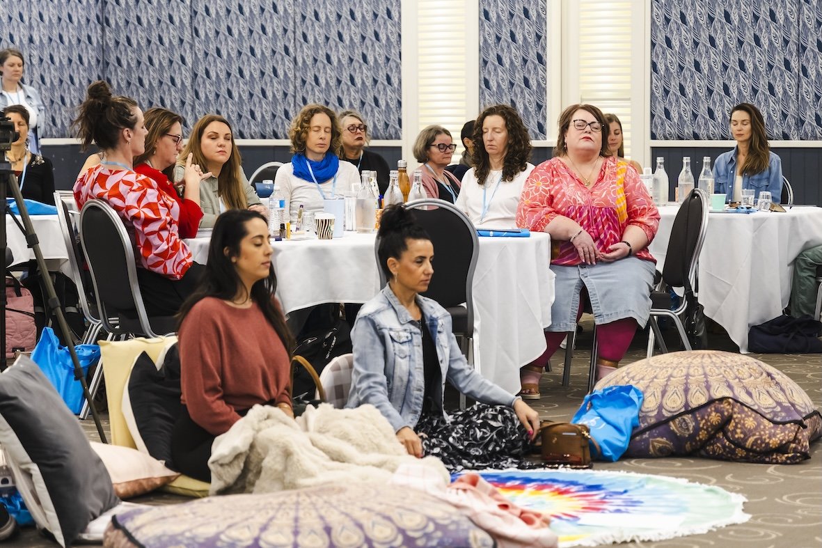 Group of women attending a meditation or yoga session in a conference room, seated cross-legged on the floor and at tables, with closed eyes and relaxed postures.