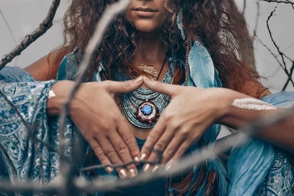 Woman dressed in shall and feathers wearing a large necklace while holding her hands in a v shape around her heart.