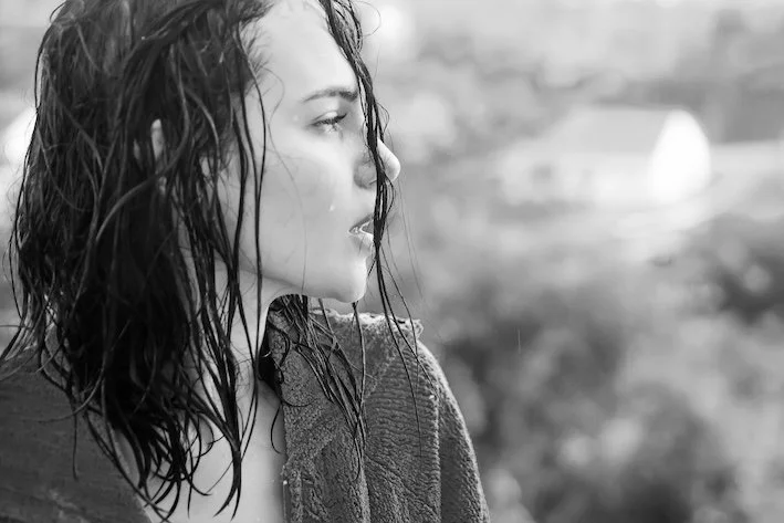 Woman in rain with wet hair and face looking off camera in a faded background.