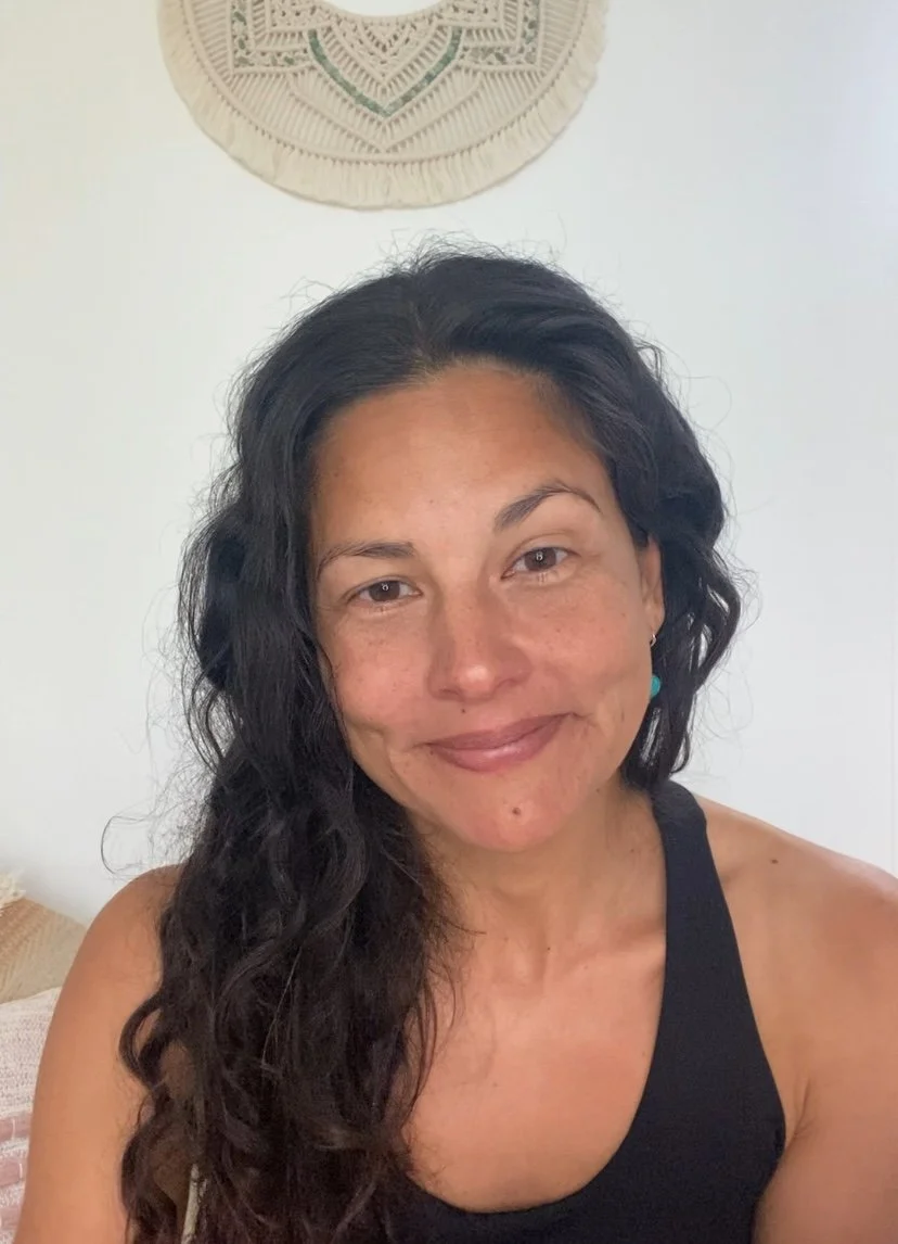 A woman with long, wavy dark hair smiling in a room with light-colored walls and a decorative wall hanging above her head.
