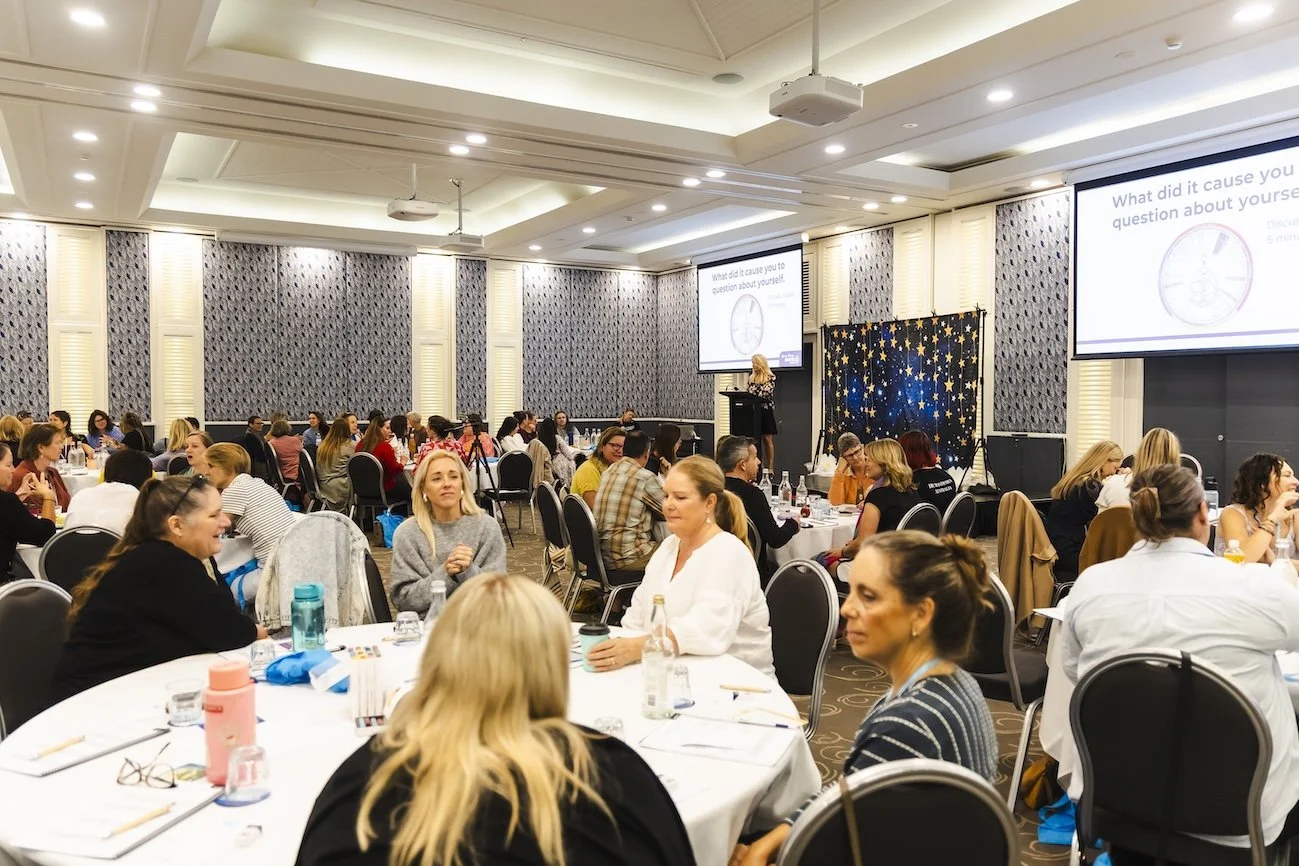 Conference room filled with women seated at round tables, attending a presentation with a speaker on stage and two large screens displaying a question, in a hotel ballroom with elegant decor.