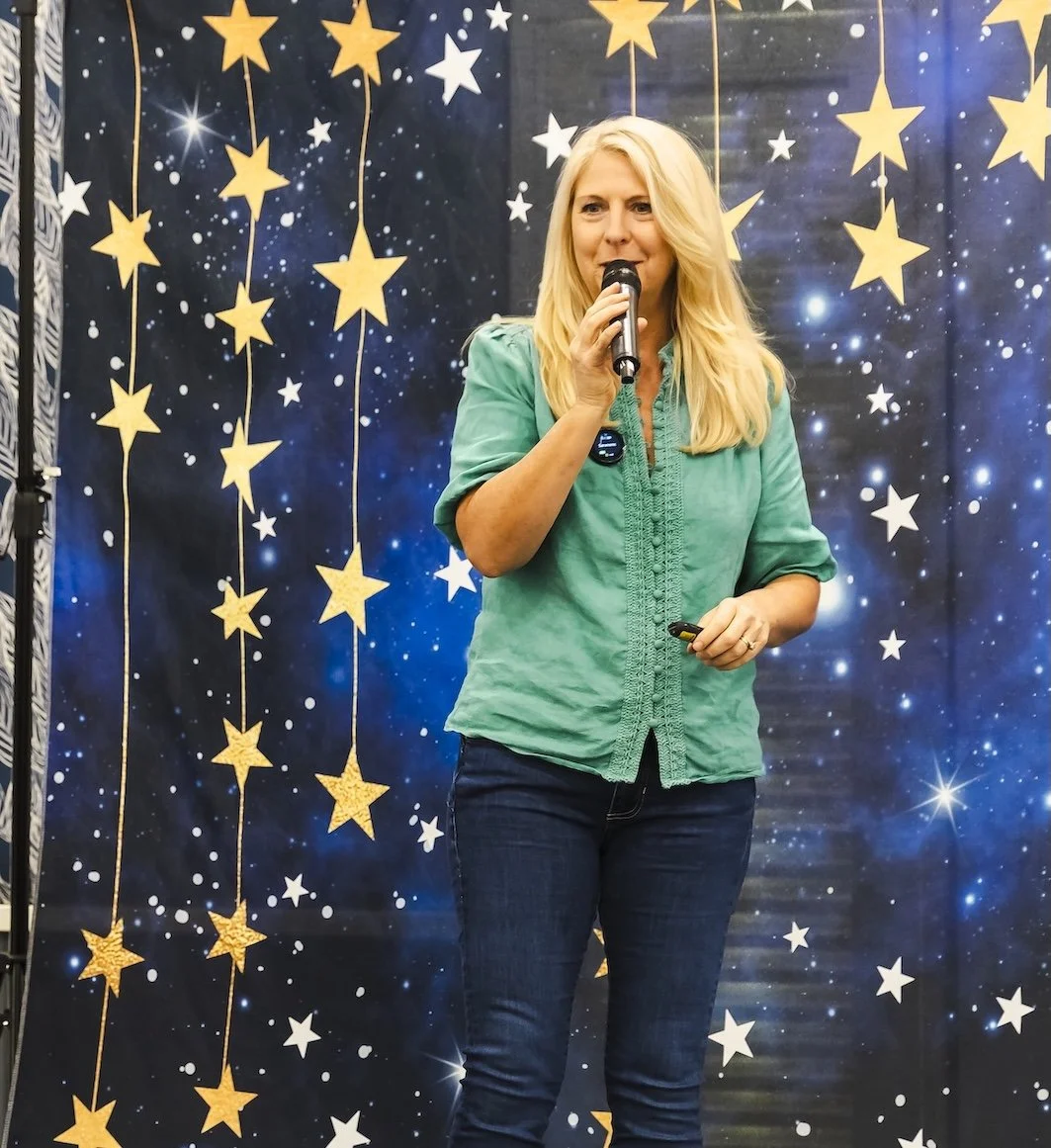 International Speaker Amber Clements in green blouse speaking into a microphone at the human design conference against a starry background with hanging yellow stars.