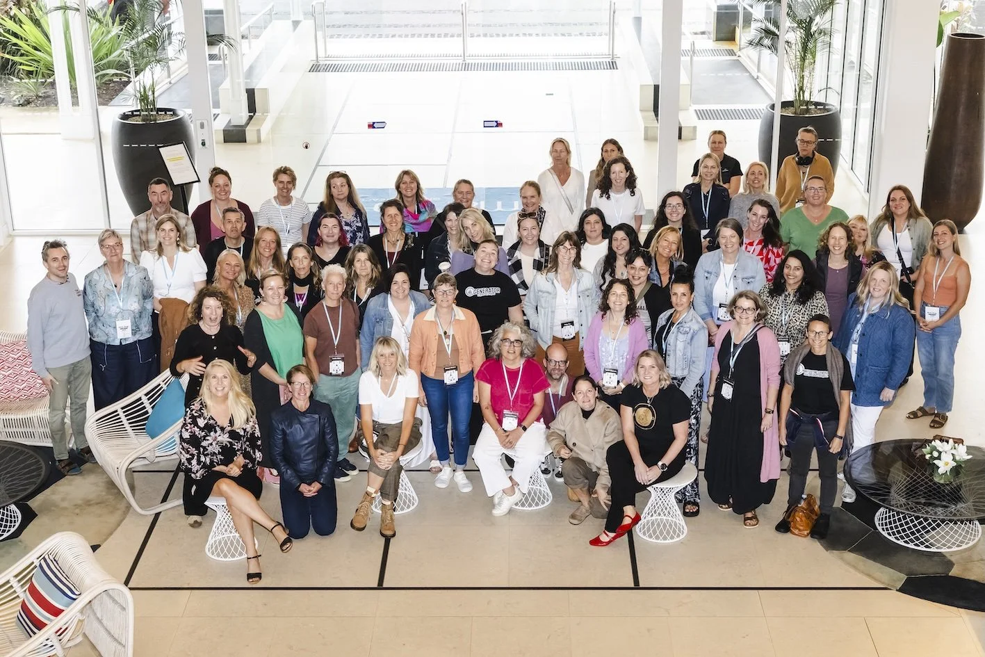 A large group of people gathered inside a modern, bright lobby with large windows and potted plants, posing for a group photo.