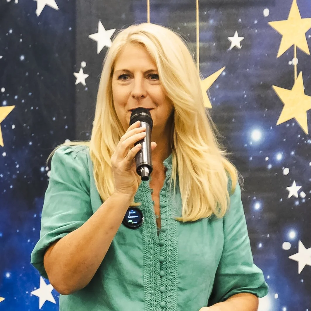 Amber Clements holding a microphone in front of a backdrop decorated with stars speaking at a human design event.