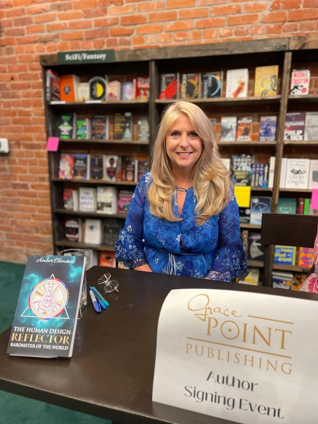 Amber Clements wearing a blue floral dress, sitting at a table with books and pens in front of her, smiling at a book signing event.