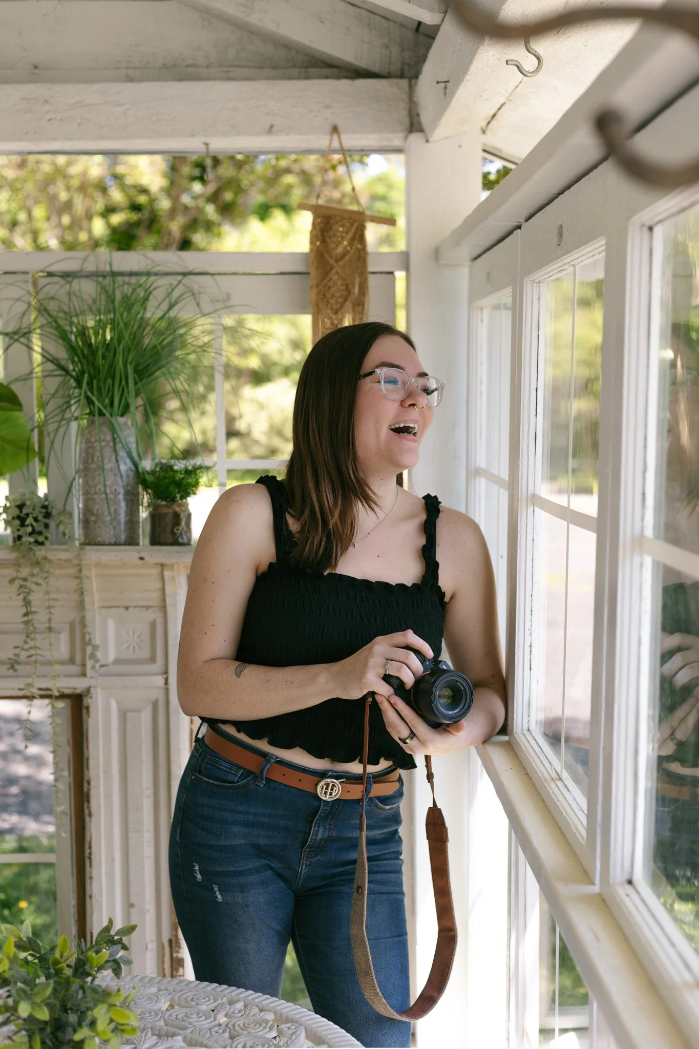 A young woman with glasses, dark hair, wearing a black sleeveless top and blue jeans, holding a camera, looking out a large window with a smile.