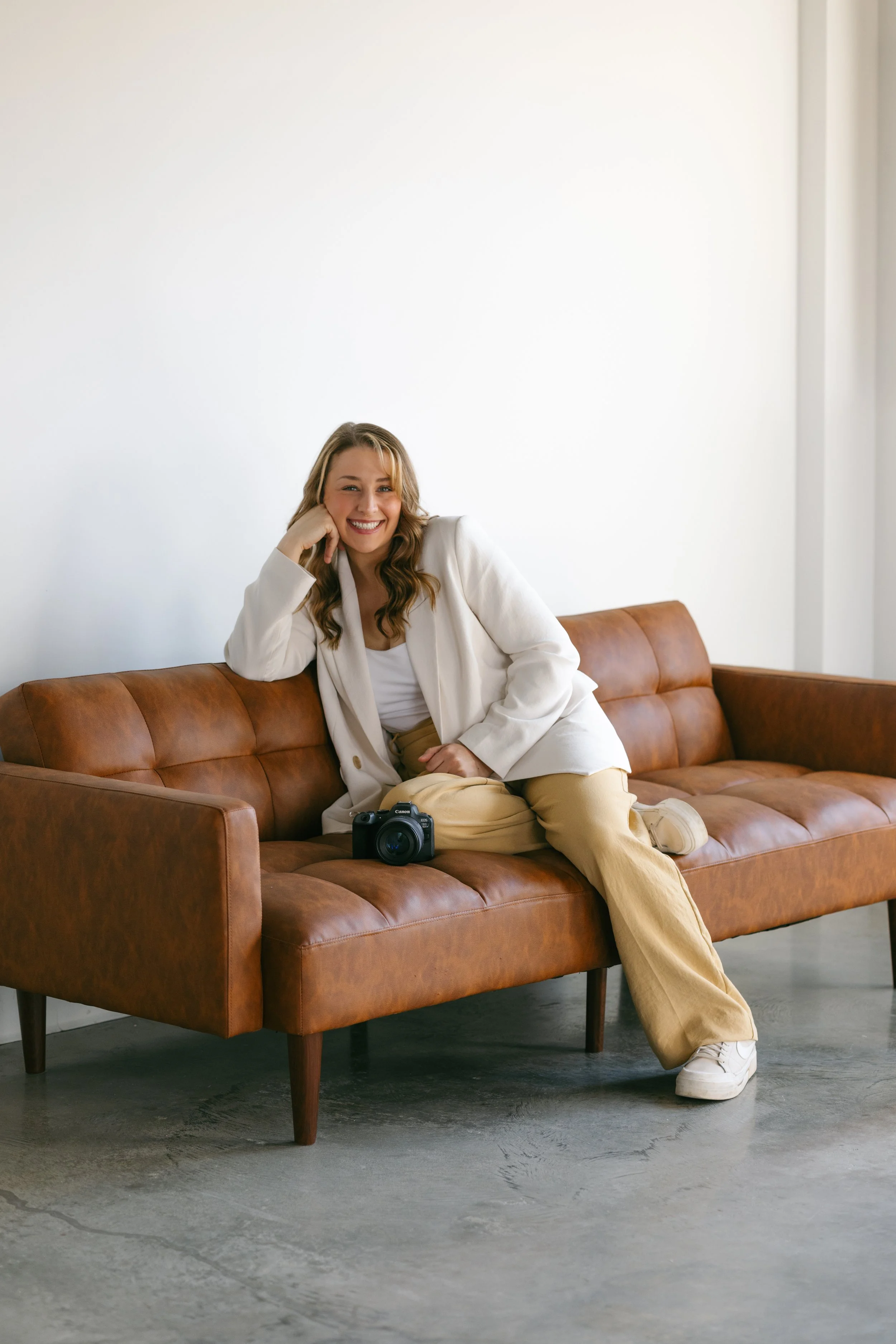 Smiling woman sitting on a brown leather sofa with a camera on her lap in a minimalist room with a white wall.