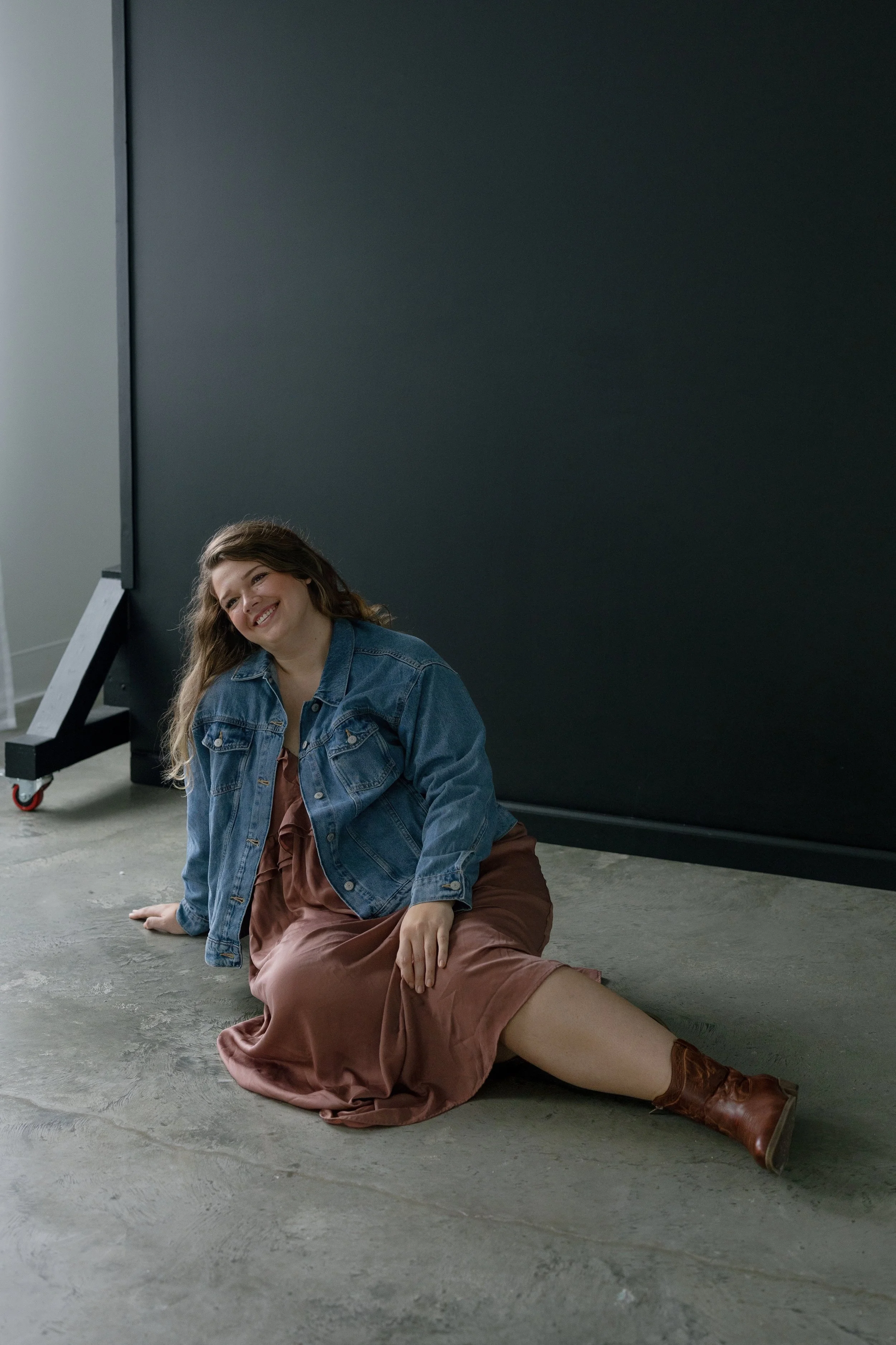 A woman in a brown dress, denim jacket, and brown boots sitting on the floor, smiling, against a dark wall in a studio setting.