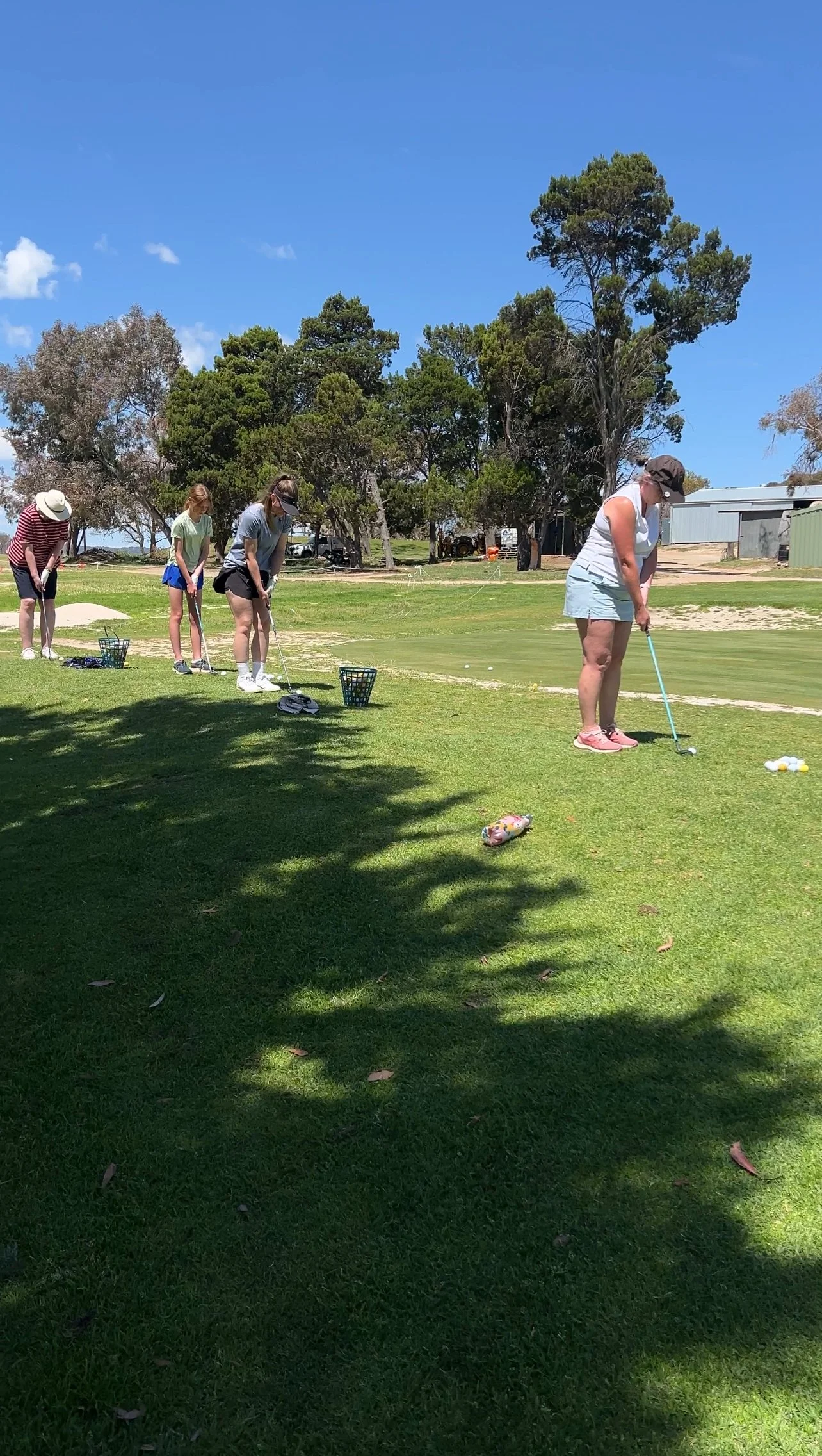 Four women practicing golf on a sunny day at a golf course, standing in a line near the green, with trees in the background.