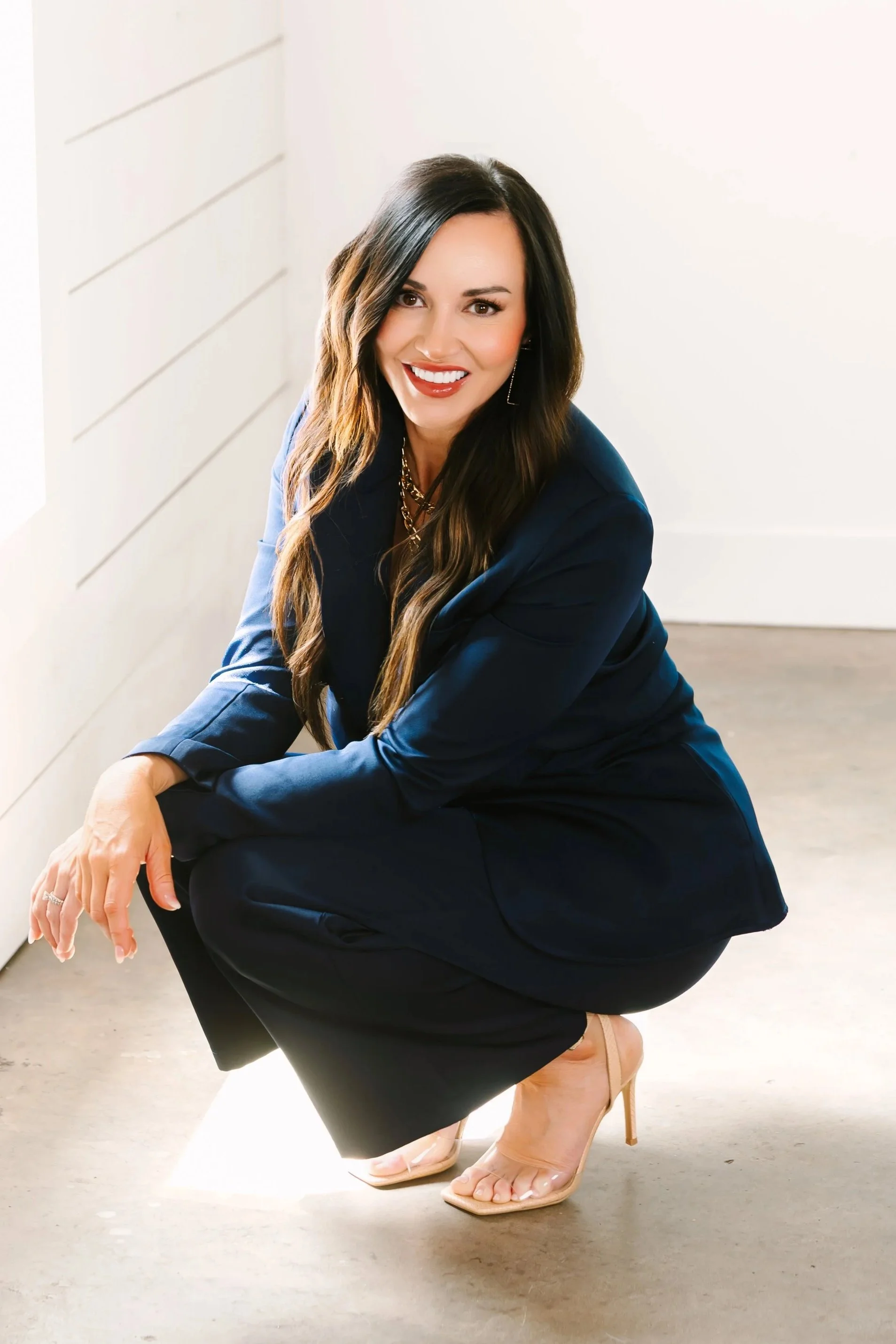 A woman with long brown hair, wearing a black shirt and ripped jeans, sitting on a white stool in front of a dark blue cabinet with shelves containing glass jars, containers, and a framed photo.