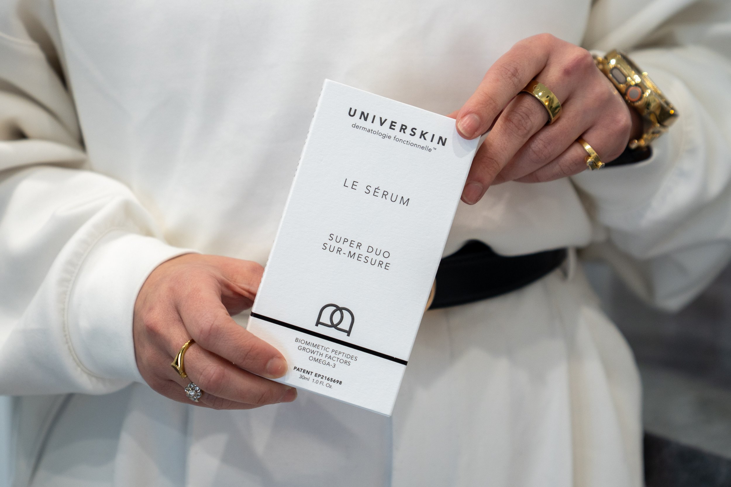 Person holding a skincare product packaging labeled 'UNIVERSKIN'. The packaging mentions skincare ingredients and benefits, including 'gland, growth factors, omega-3'. The person is wearing jewelry, including a gold ring and a bracelet, and is dressed in a light-colored garment.