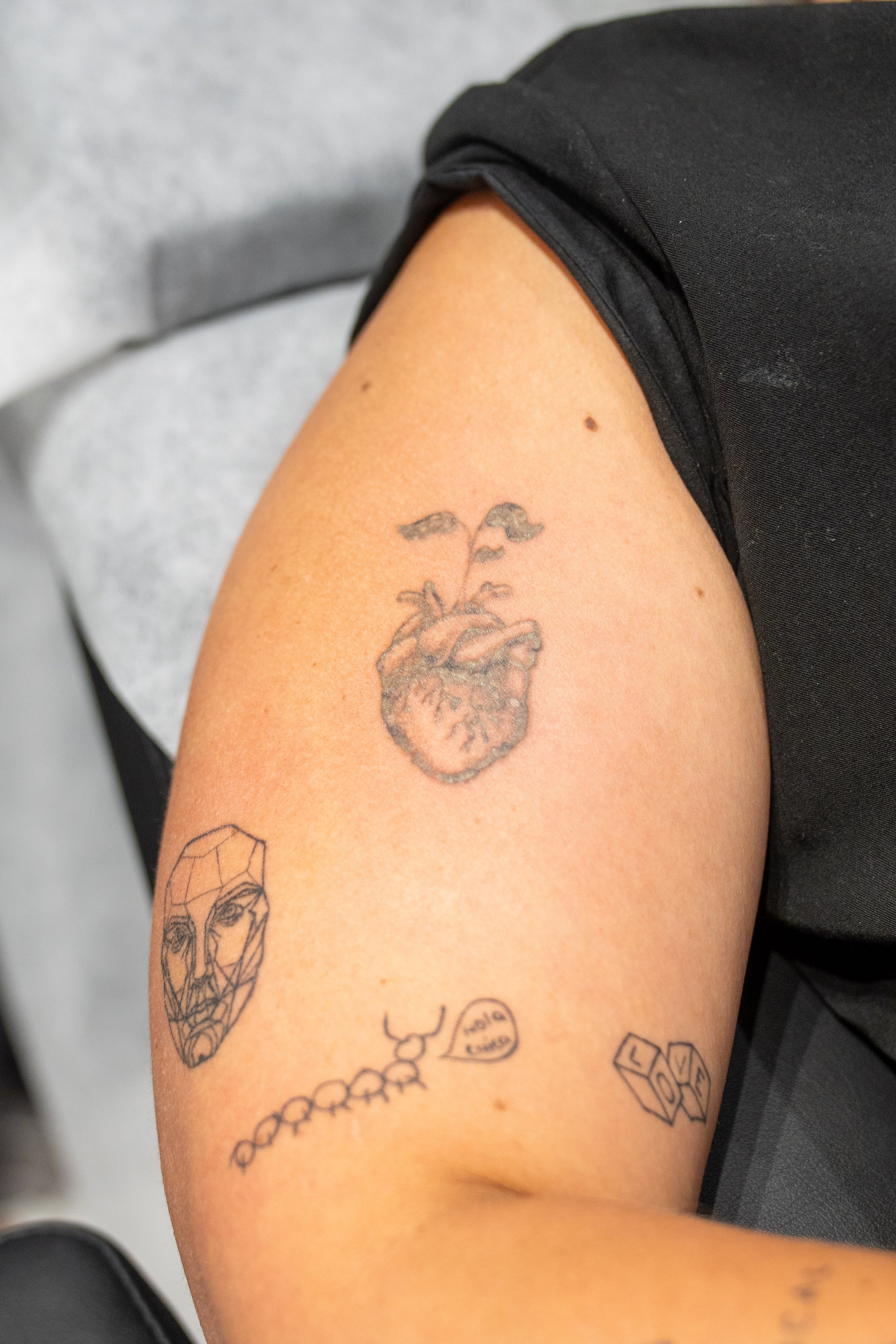 Close-up of a person's upper arm with multiple tattoos, including an anatomical heart, a face within a geometrical shape, a string of beads, a speech bubble, and some text, with the person wearing a black shirt and sitting against a gray background.