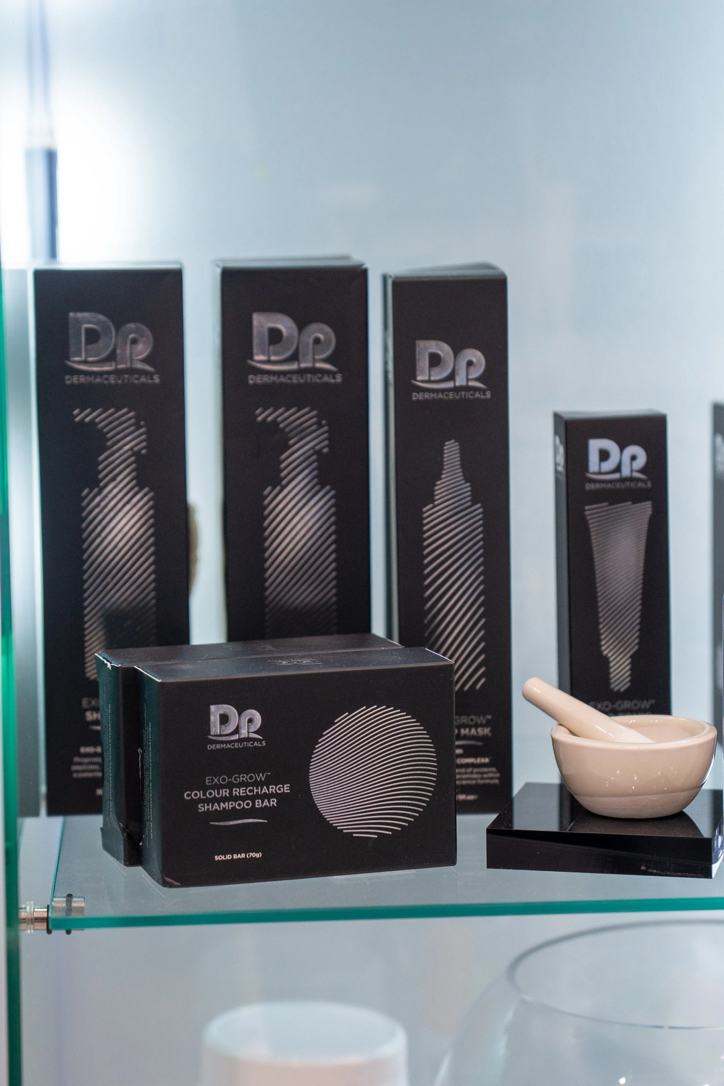 A glass display case containing skincare products, including a black box labeled 'EXO-GROW Colour Recharge Shampoo Bar' and several similar black boxes in the background with the branding 'Dermo Cosmetics' and stylized logo.