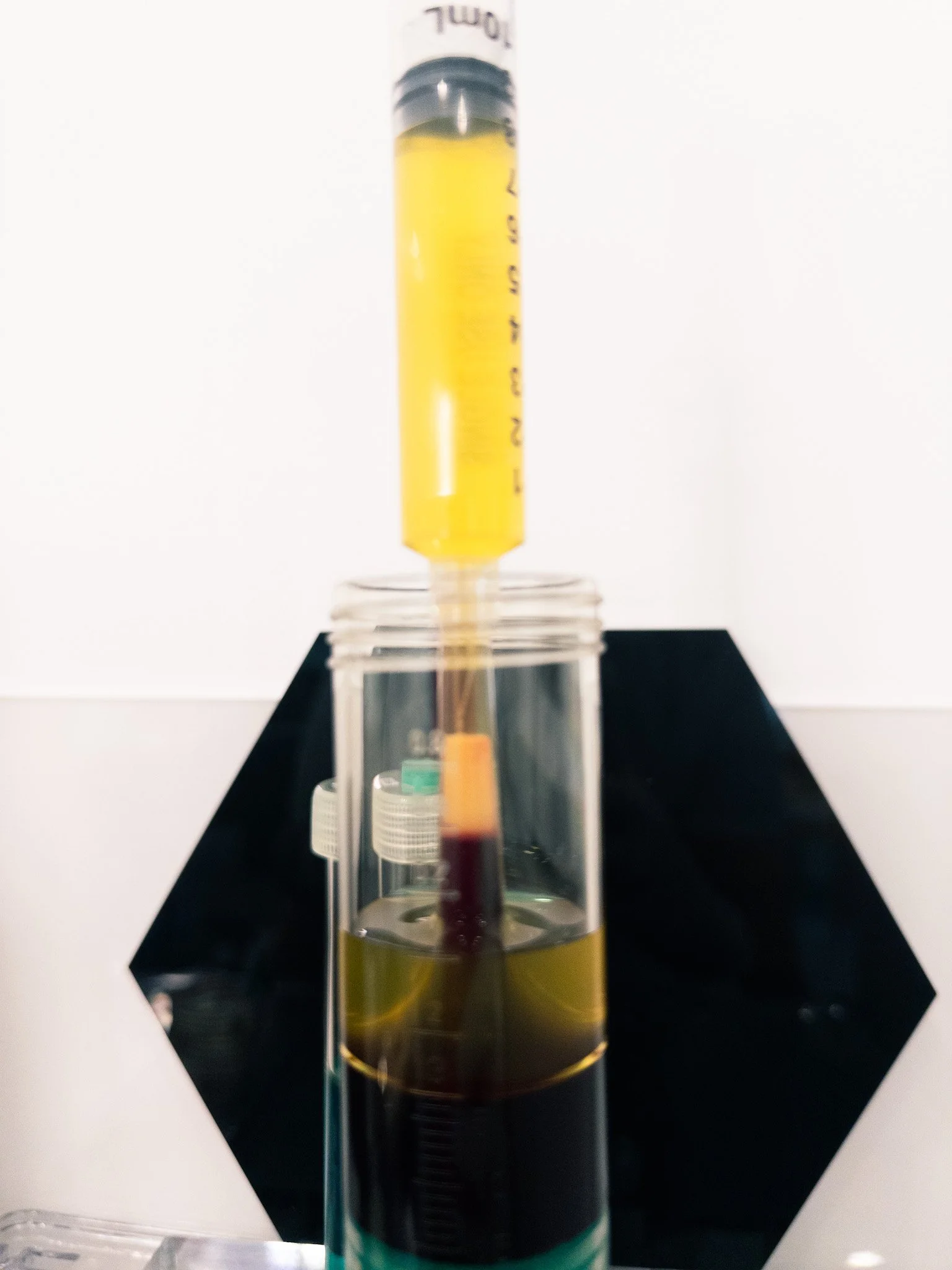 Close-up view of a syringe with yellow liquid, positioned vertically on a black platform against a plain white background.
