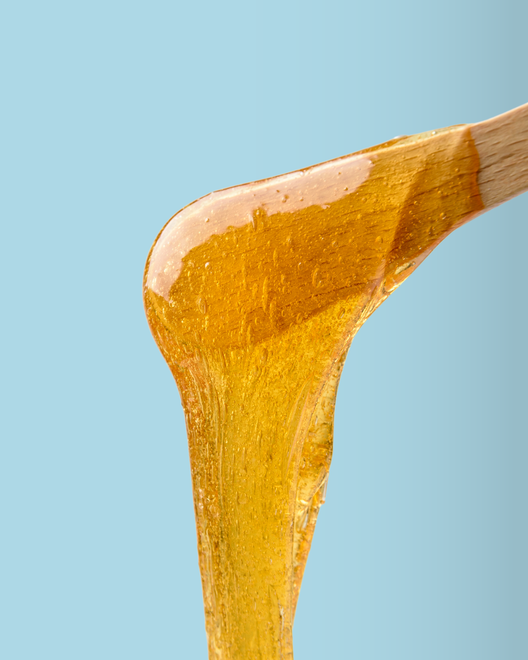 Close-up of golden honey being on a wooden honey dipper against a light blue background.