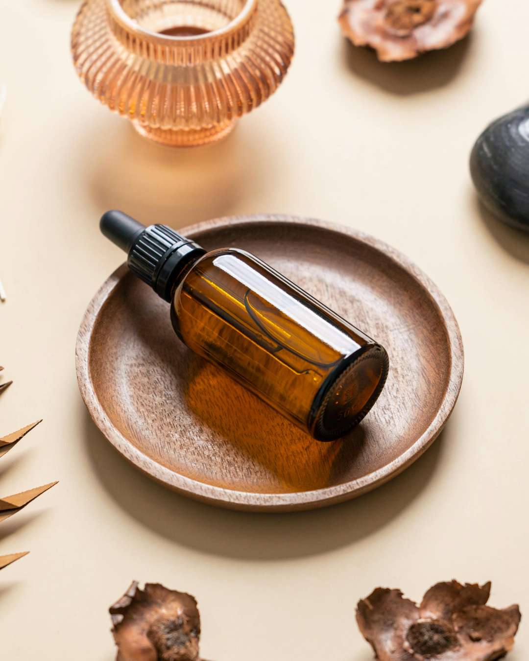 An amber glass dropper bottle resting on a round wooden dish, surrounded by decorative objects including a pink and gold jewelry holder, a black stone, and dried plant elements on a light-colored surface.