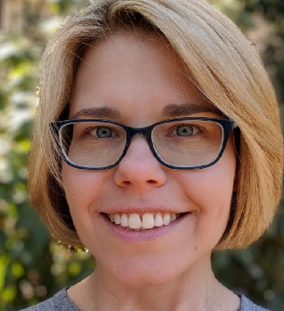 A woman with shoulder-length blonde hair, wearing glasses, smiling outdoors with greenery in the background.