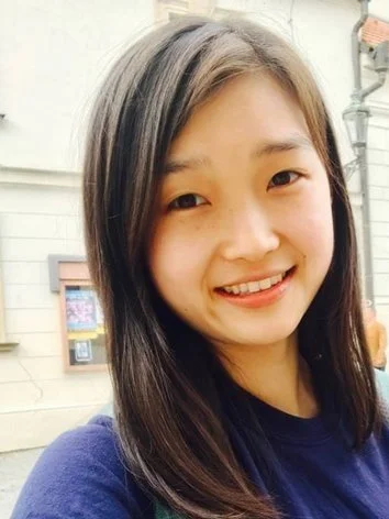 A young girl smiling outdoors with a light-colored building in the background.