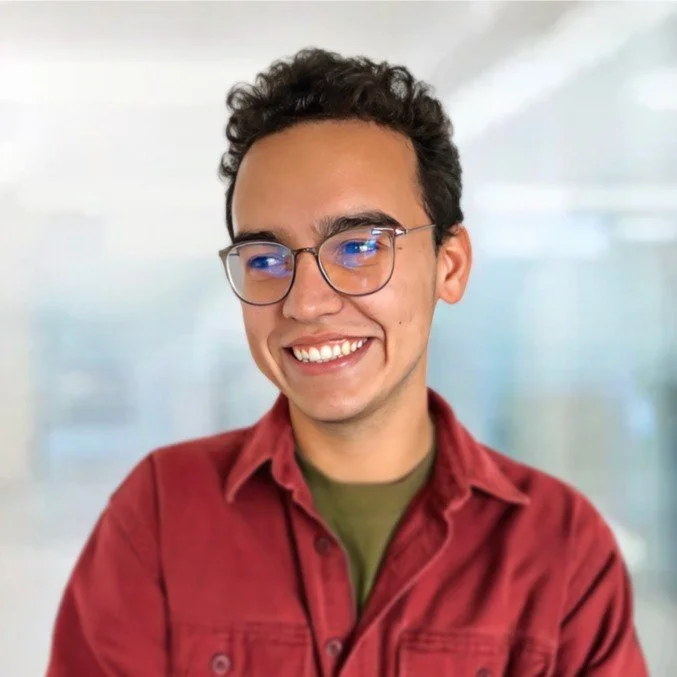 A young man with curly hair, glasses, and a bright smile wearing a red jacket over a green shirt, standing in a modern, blurred indoor background.