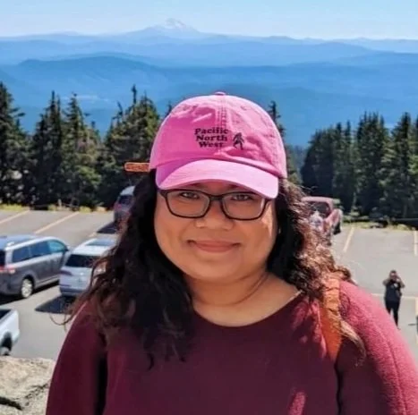 Woman wearing glasses and a pink baseball cap with mountains and trees in the background.